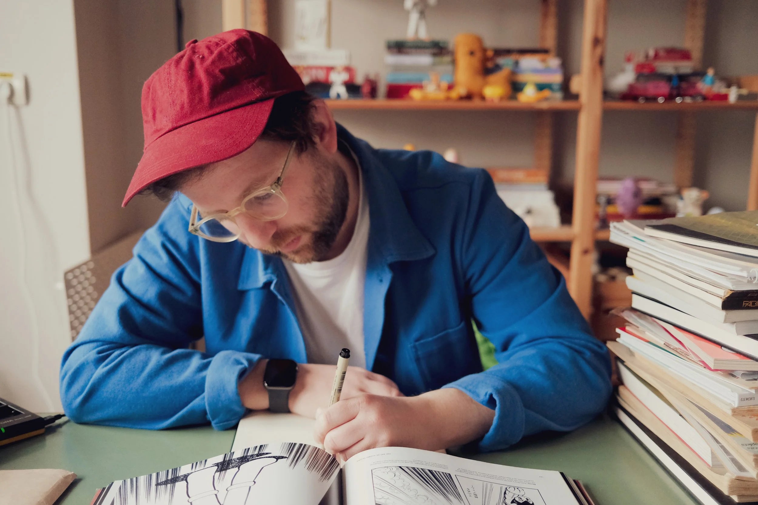 Person wearing a red cap and blue jacket sitting at a desk with books and a notebook.